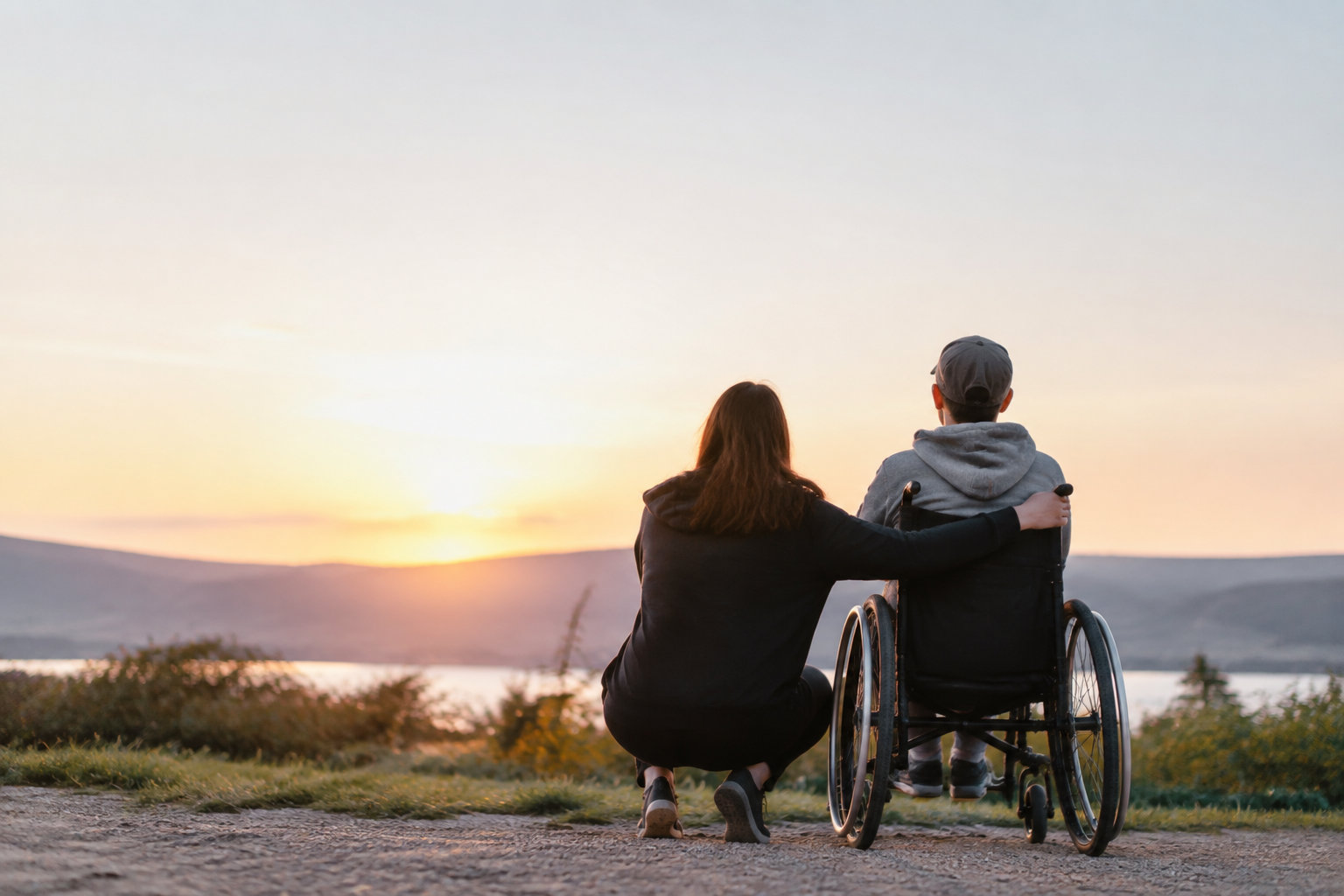 Une femme soutient une personne en fauteuil roulant face au soleil couchant sur un lac.