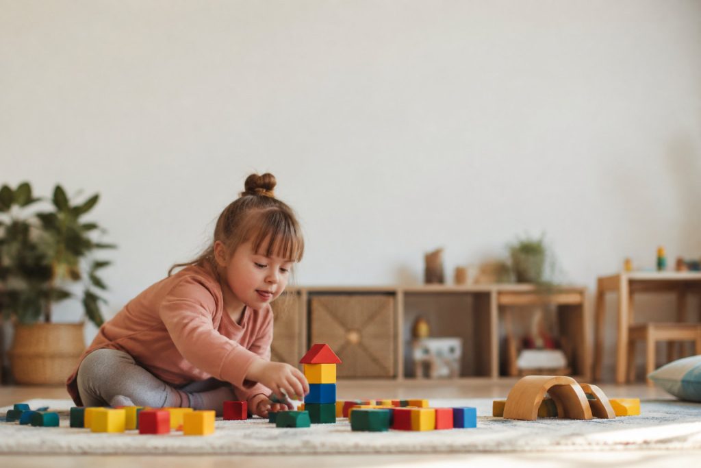 Une petite fille souriante avec trisomie 21 est assise sur un tapis et joue avec des blocs de construction colorés.
